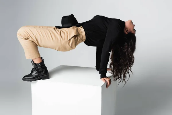 Full length of stylish model with fedora hat on beige pants posing on white cube on grey — Stock Photo