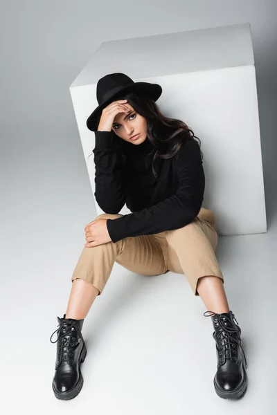 Full length of trendy model in fedora hat looking at camera while sitting near white cube on grey — Stock Photo