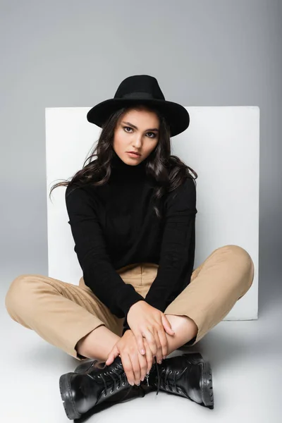 Full length of stylish model in fedora hat sitting with crossed legs near white cube on grey — Stock Photo