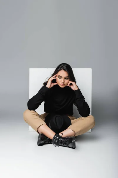 Modèle élégant en col roulé noir avec chapeau fedora assis près du cube blanc sur gris — Photo de stock