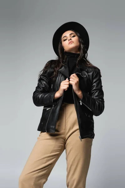 Low angle view of trendy woman in fedora hat and black leather jacket looking at camera while posing isolated on grey — Stock Photo