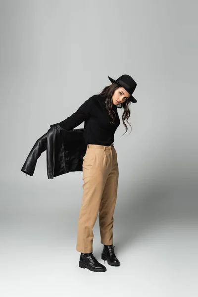 Full length of stylish woman in fedora hat holding black leather jacket while posing on grey — Stock Photo