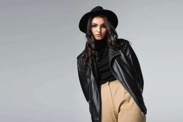 Mujer joven en sombrero fedora y chaqueta de cuero de moda posando aislado en gris - foto de stock