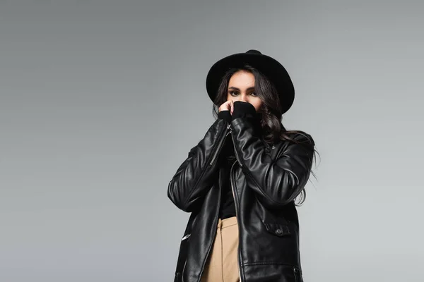 Jeune femme en chapeau de fedora et veste en cuir tendance couvrant le visage avec les mains isolées sur gris — Photo de stock