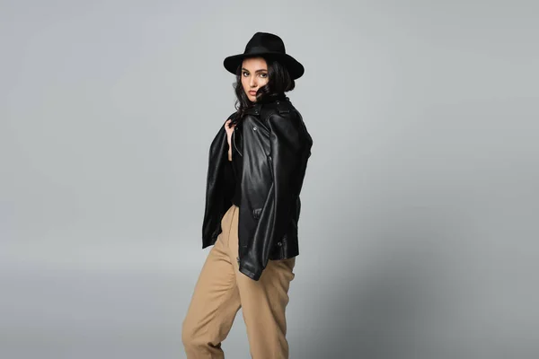 Young brunette woman in fedora hat and trendy leather jacket posing on grey — Stock Photo