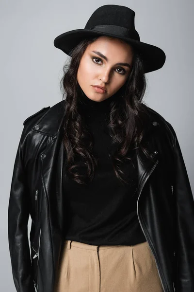 Young brunette woman in fedora hat, black turtleneck and trendy leather jacket posing isolated on grey — Stock Photo