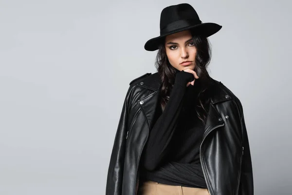 Молодая женщина в шляпе-шляпе, черной водолазке и модной кожаной куртке позирует изолированно на сером — стоковое фото