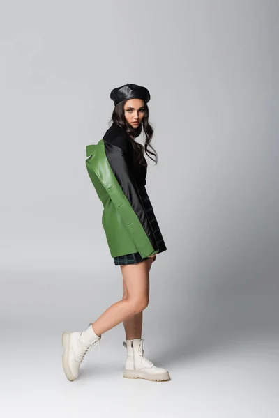 Повна довжина молодої модної жінки в береті і зеленій шкіряній куртці позує на сірому — стокове фото