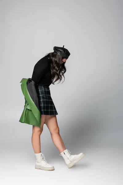 Volle Länge der jungen trendigen Frau in Baskenmütze, kariertem Rock und grüner Lederjacke posiert auf grau — Stockfoto
