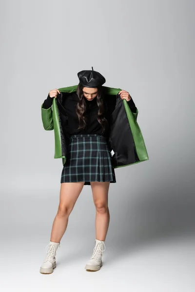 Pleine longueur de jeune femme élégante en béret et jupe à carreaux ajustant veste en cuir vert sur gris — Photo de stock