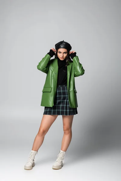 Full length of young stylish woman plaid skirt and green leather jacket adjusting beret on grey — Stock Photo