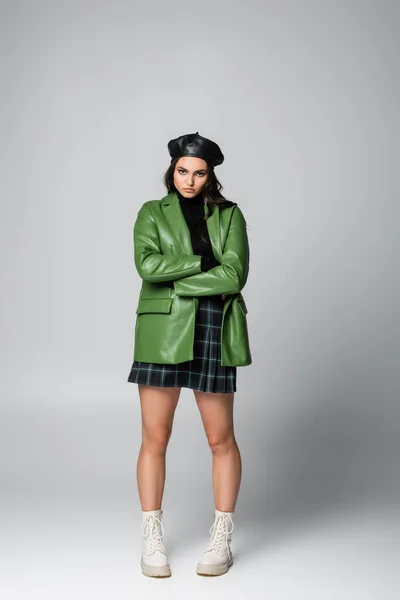 Pleine longueur de jeune femme élégante en béret, jupe à carreaux et veste en cuir vert posant avec les bras croisés sur gris — Photo de stock