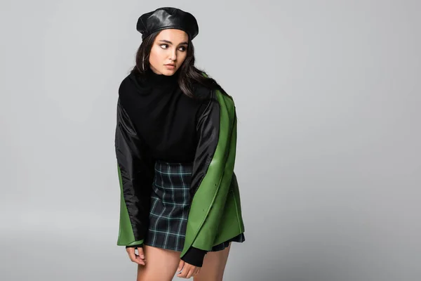 Junge trendige Frau in Baskenmütze, kariertem Rock und grüner Lederjacke posiert isoliert auf grau — Stockfoto