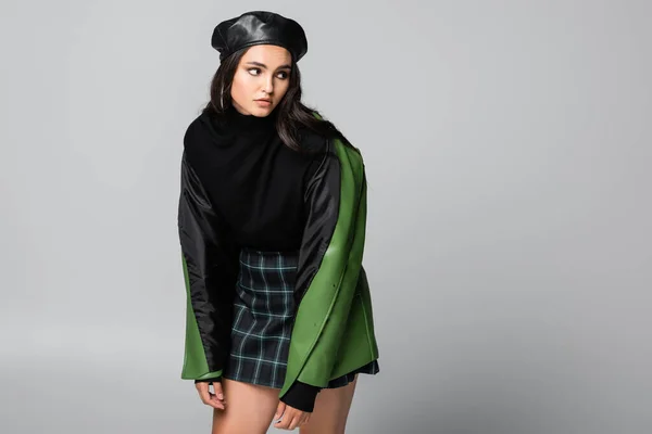 Mujer joven de moda en boina, falda a cuadros y chaqueta de cuero verde posando aislado en gris - foto de stock