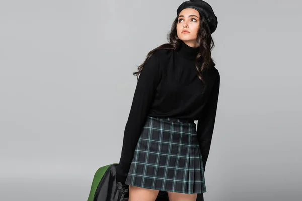 Young trendy woman in beret and plaid skirt holding green leather jacket isolated on grey — Stock Photo