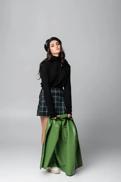 Full length of young stylish woman in beret and plaid skirt holding green leather jacket on grey — Stock Photo