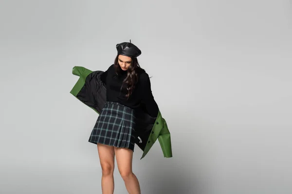 Young stylish woman in plaid skirt, beret and green jacket posing on grey — Stock Photo