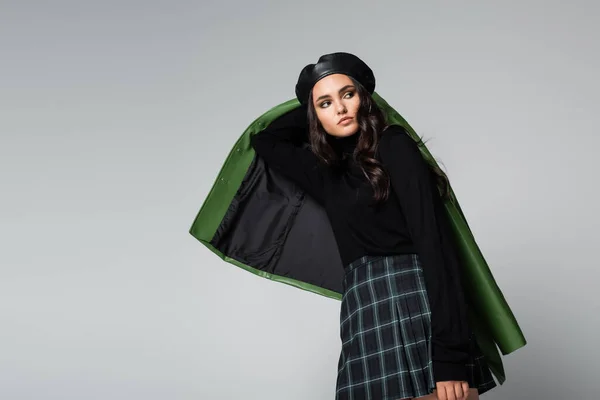 Young brunette woman in plaid skirt, black beret and jacket posing isolated on grey — Stock Photo