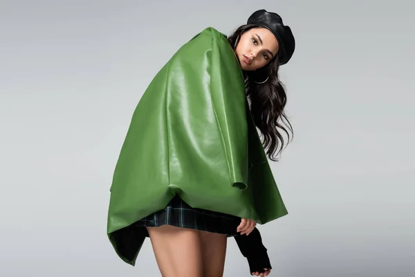 Junge stylische Frau in schwarzem Baskenmütze und grüner Lederjacke posiert isoliert auf grau — Stockfoto