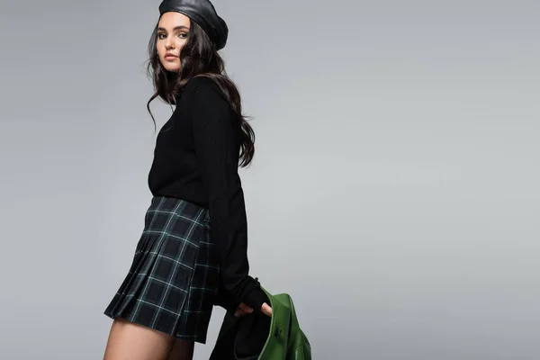 Mujer elegante en boina sosteniendo chaqueta de cuero verde mientras posando aislado en gris - foto de stock