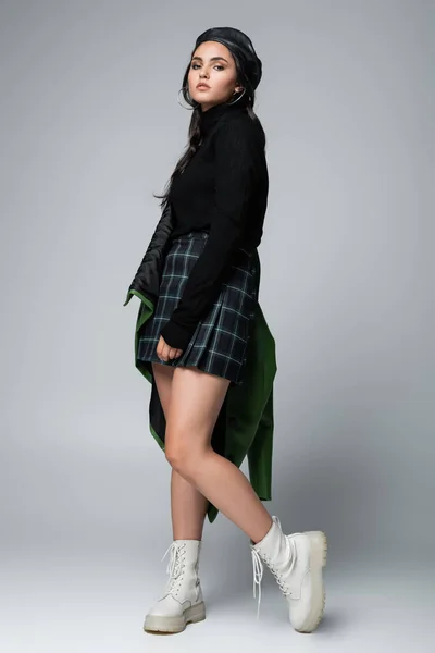 Comprimento total da jovem mulher em jaqueta de couro verde, saia quadriculada e boina preta posando no cinza — Fotografia de Stock
