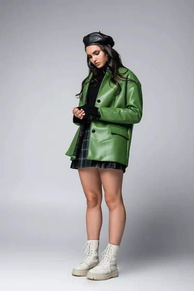 Full length of young model in green jacket, plaid skirt and black beret posing on grey — Stock Photo