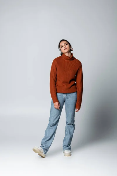 Full length of young woman in sweater and denim jeans on grey — Stock Photo