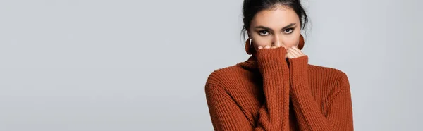 Young woman in knitted sweater looking at camera while covering face isolated on grey, banner — Stock Photo