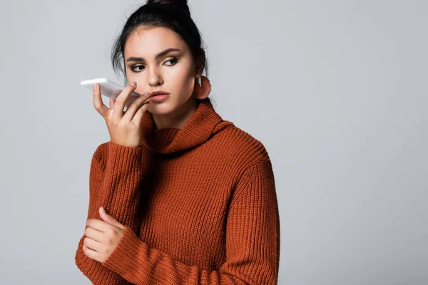 Молодая женщина в трикотажном свитере записывает голосовое сообщение на смартфон, изолированный на сером — стоковое фото
