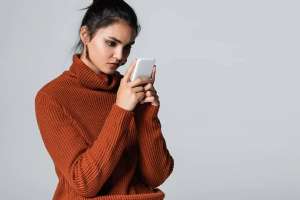 Junge Frau in Strickpullover-Botschaft auf Smartphone isoliert auf grau — Stockfoto