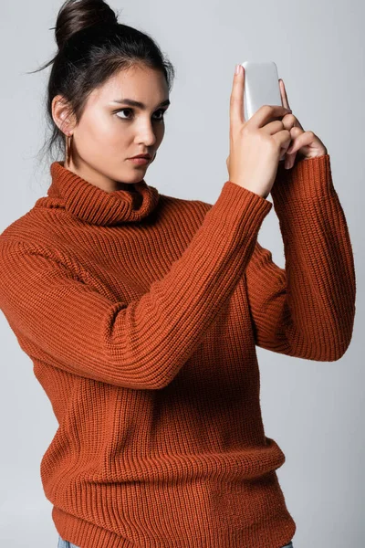 Giovane donna in maglione lavorato a maglia con smartphone isolato su grigio — Foto stock