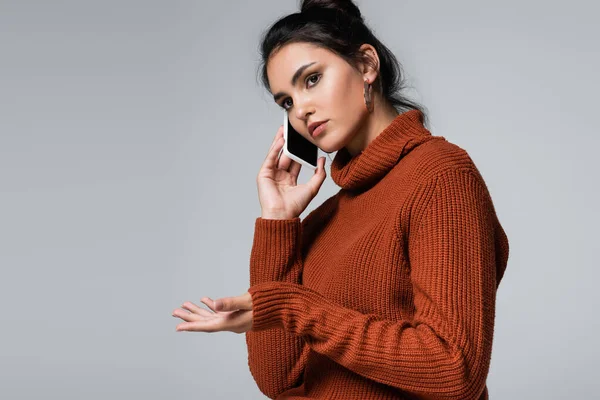 Молода жінка в трикотажному светрі говорить на смартфоні, жестикулюючи ізольовано на сірому — стокове фото
