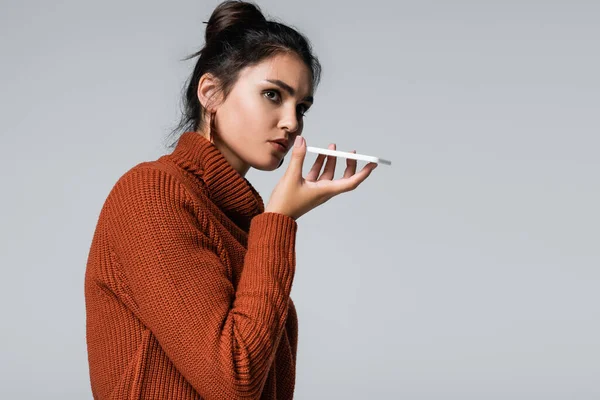 Young woman in warm sweater recording voice message on smartphone isolated on grey — Stock Photo