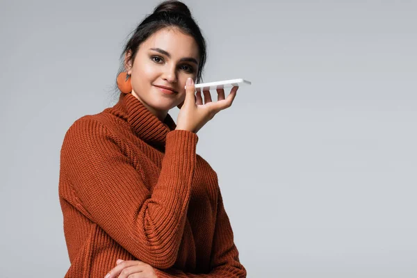 Glückliche junge Frau im Strickpullover, die Sprachnachricht auf dem Handy aufnimmt, isoliert auf grau — Stockfoto