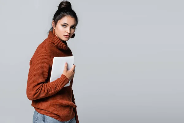 Morena mujer joven en jersey de punto sosteniendo tableta digital aislado en gris - foto de stock