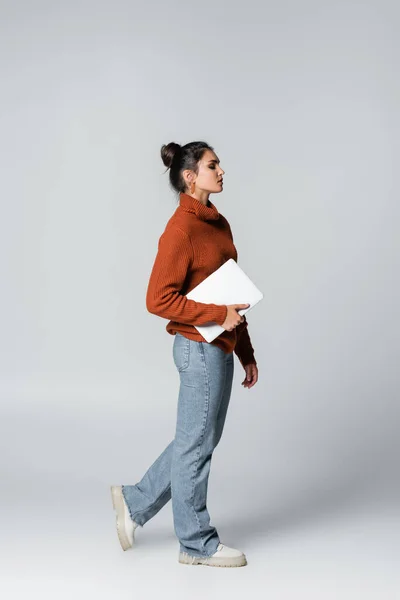 Longitud completa de la mujer joven en suéter de punto y pantalones vaqueros celebración portátil en gris - foto de stock