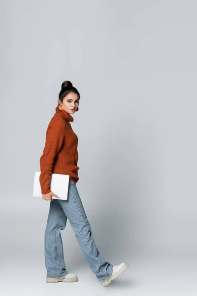 Longitud completa de la mujer joven en suéter de punto y pantalones vaqueros que sostienen el ordenador portátil y caminar sobre gris - foto de stock