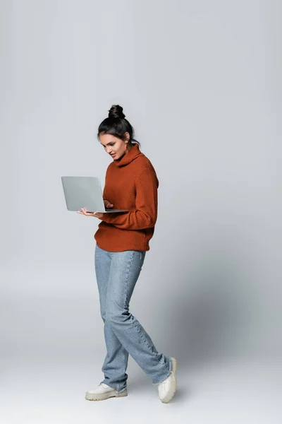 Повна довжина молодої жінки в трикотажному светрі і джинсах, використовуючи ноутбук на сірому — стокове фото