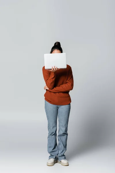 Full length of young freelancer in knitted sweater and jeans covering face while using laptop on grey — Stock Photo