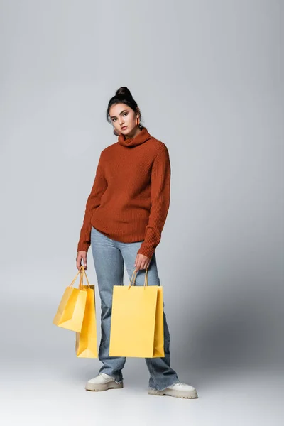 Full length of young woman in sweater and denim jeans holding yellow shopping bags on grey — Stock Photo