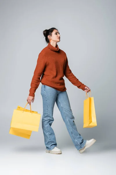 Full length of young model in sweater and jeans holding yellow shopping bags on grey — Stock Photo