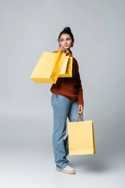 Full length of young shopaholic in sweater and jeans holding yellow shopping bags on grey — Stock Photo