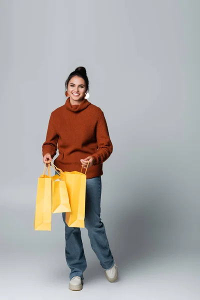 Full length of joyful young woman in sweater and jeans holding yellow shopping bags on grey — Stock Photo