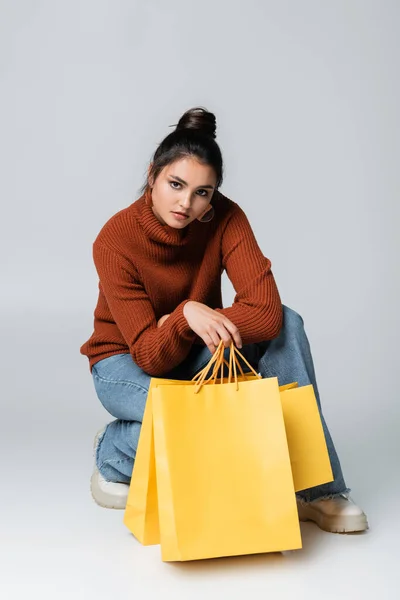 Comprimento total da jovem mulher em suéter e jeans posando perto de sacos de compras amarelos em cinza — Fotografia de Stock