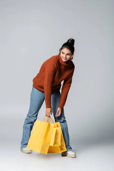 Полная длина веселой молодой женщины в свитере и джинсах, держащих желтые пакеты покупок на сером — стоковое фото