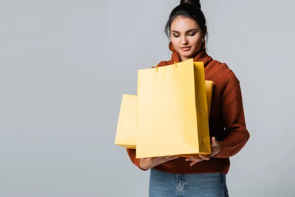 Giovane donna in maglione guardando borse della spesa gialle isolate su grigio — Foto stock