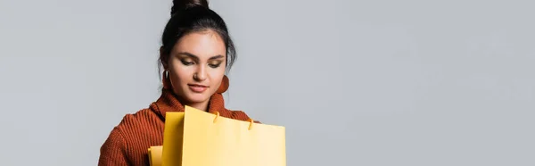 Молода жінка в светрі, дивлячись на жовту сумку для покупок ізольована на сірому, банер — стокове фото
