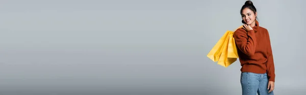 Щаслива молода жінка в светрі і джинсах тримає жовті сумки для покупок на сірому, банер — стокове фото