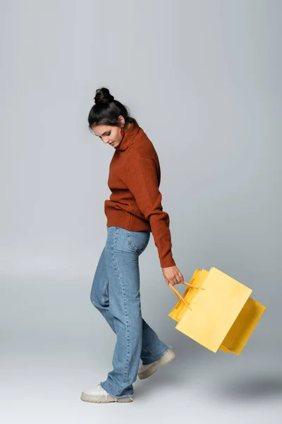 Pleine longueur de jeune femme en pull tenant des sacs à provisions jaunes et marchant sur gris — Photo de stock