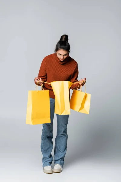 Longueur totale de la jeune femme en pull et jeans tenant des sacs à provisions jaunes sur gris — Photo de stock